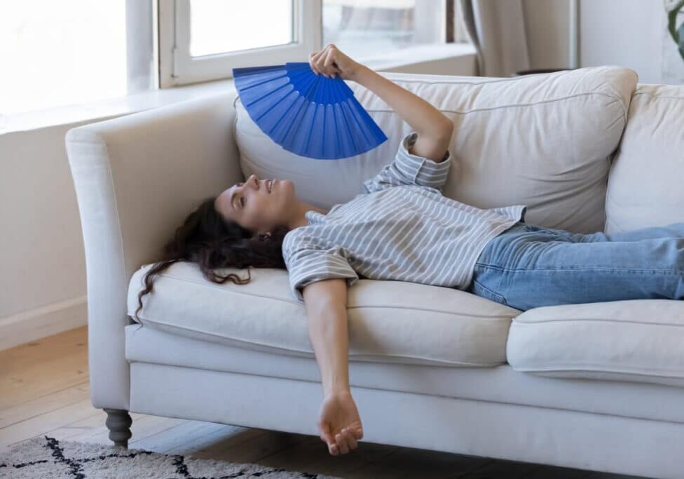 Exhausted young woman suffering from hot weather, overheat, too high air temperature at home, lying on sofa, waving paper handheld fan, cooling, feeling discomfort